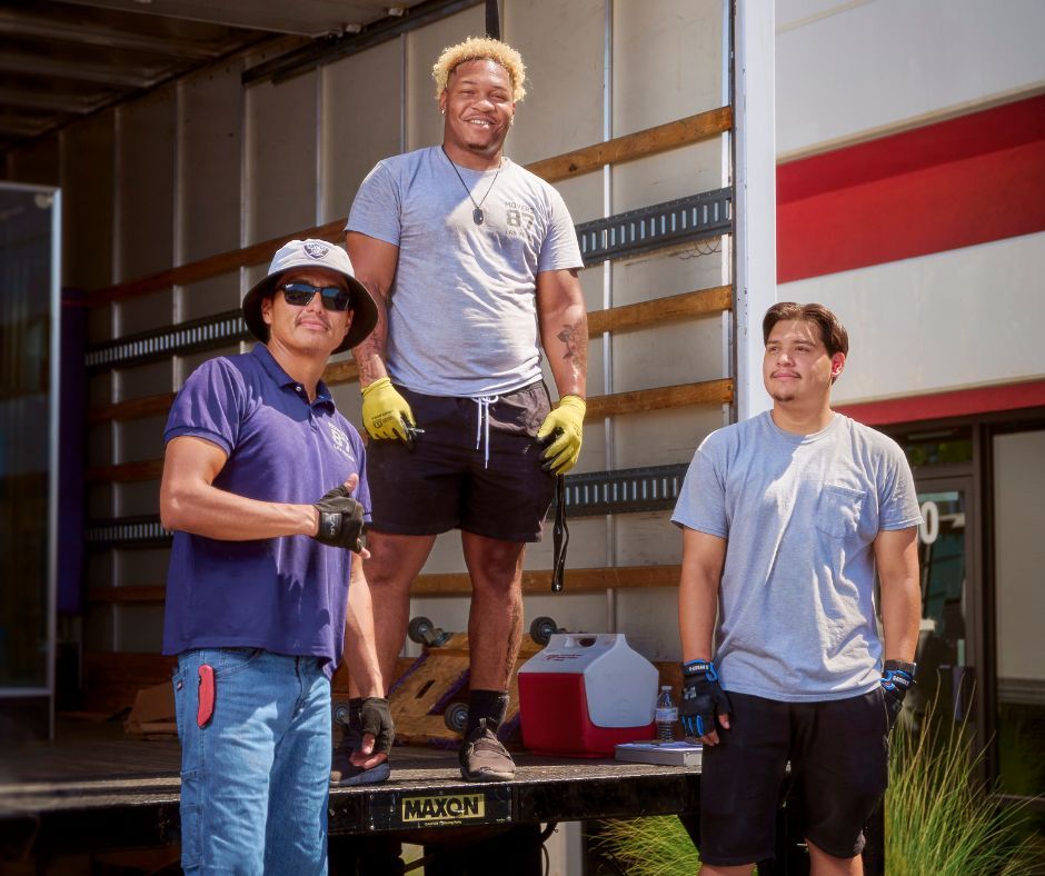 Three movers standing in front of a truck, smiling. One is wearing a hat, another is on a lift, and all are in work clothes.