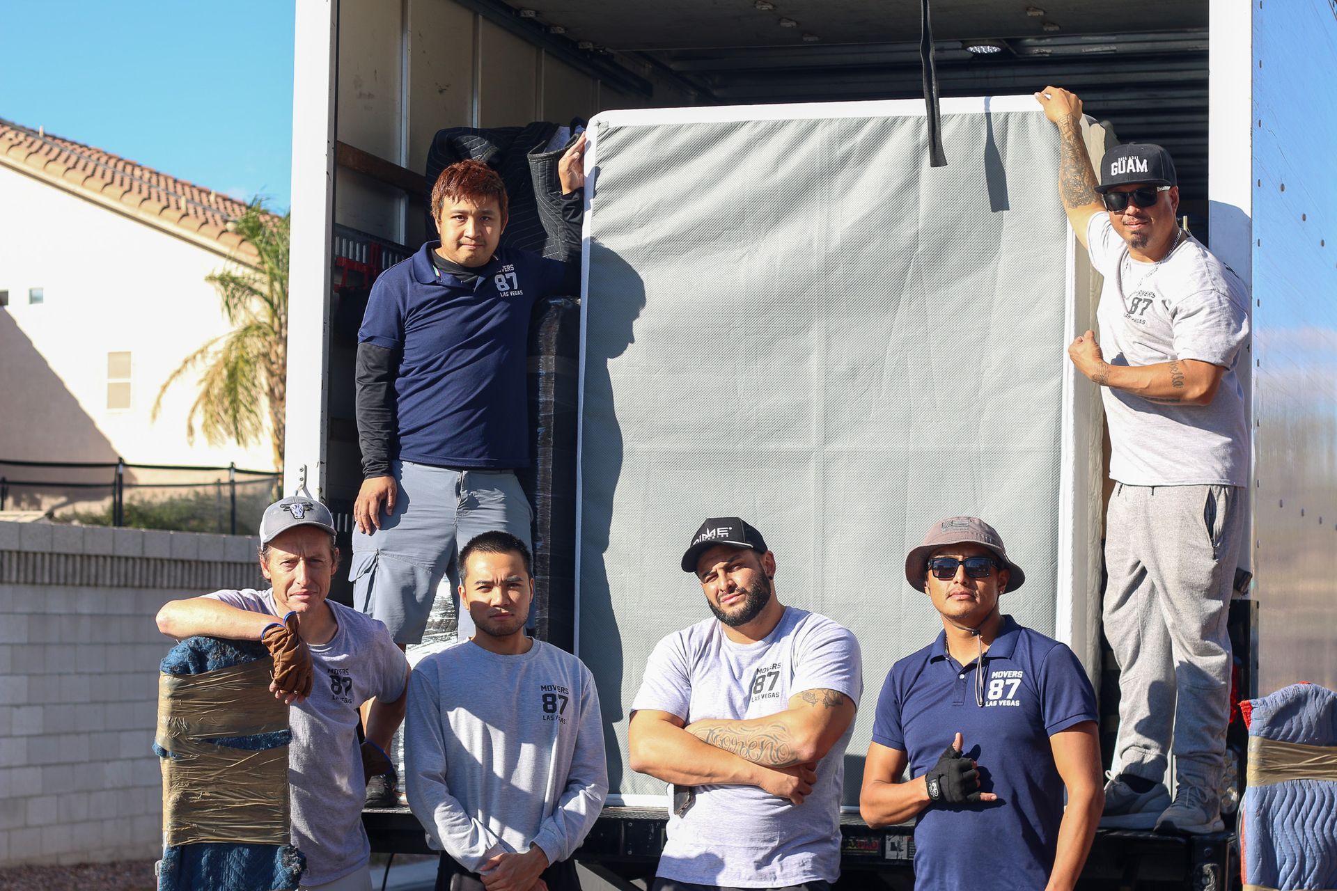Six movers standing in front of a moving truck with a large mattress, sunny outdoors.