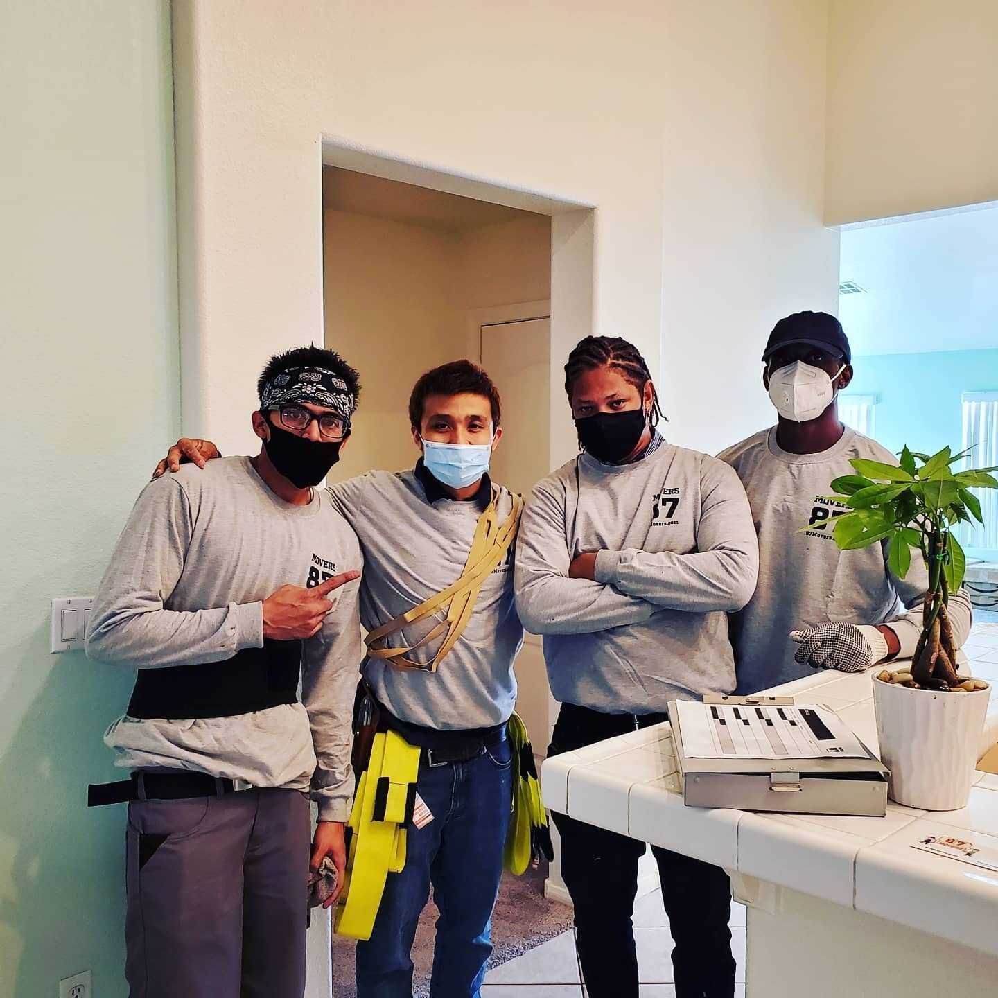 Four people in work attire pose indoors, two with masks. One has a tool belt, others have arms crossed. There's a plant and tools on a counter.
