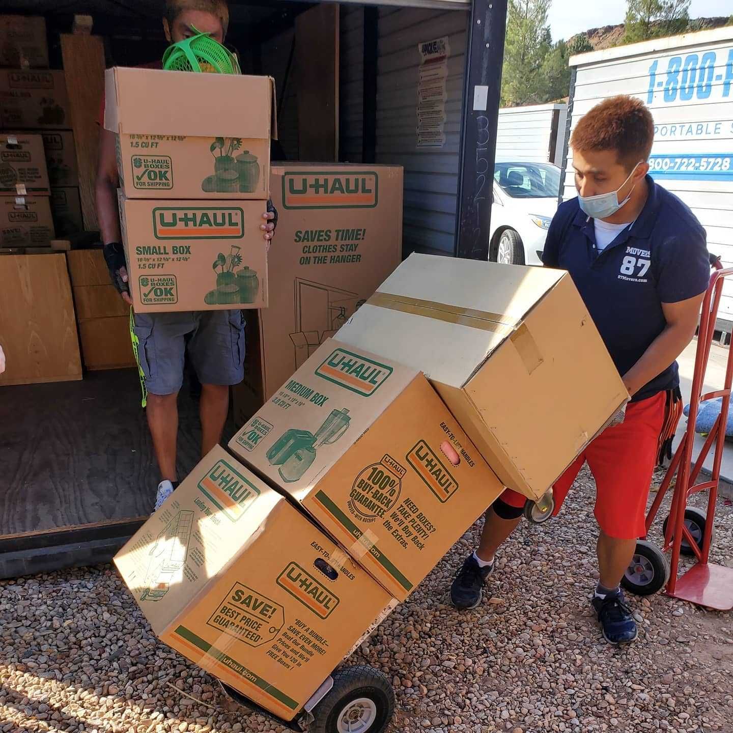 Two people loading U-Haul boxes into a storage unit; one carrying boxes, the other using a dolly.