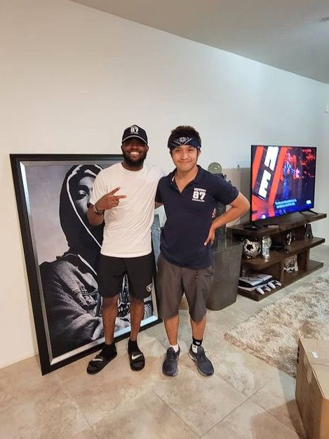 Two people stand beside a large framed portrait in a living room. One person gestures a peace sign. A TV is visible.