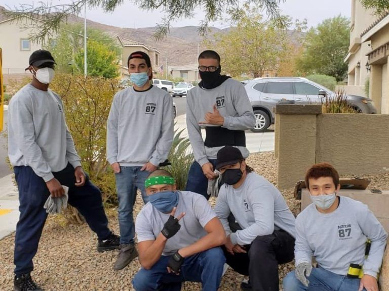 Group of people wearing masks and gray shirts pose outdoors. Some are crouching or leaning, with a residential backdrop.