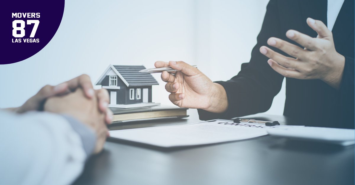 Hands reaching over paperwork, model house, pen; business setting.