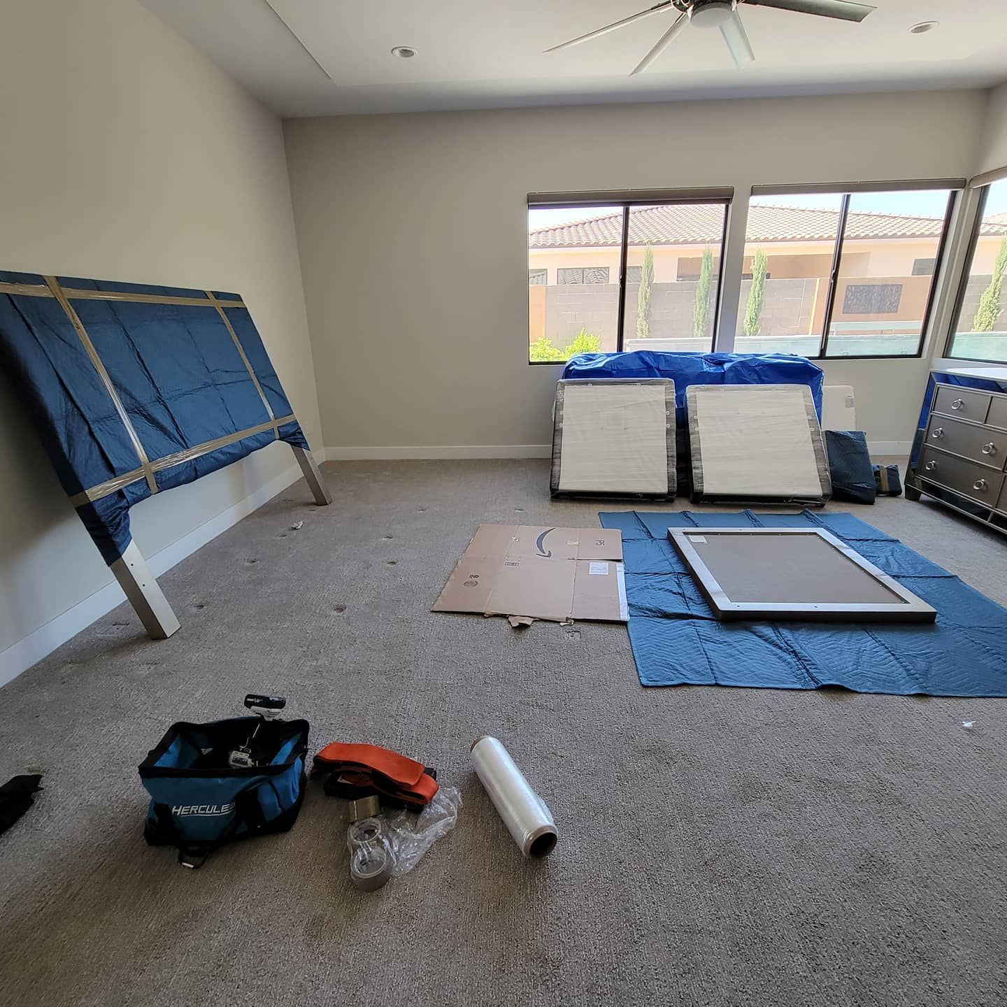 Empty room with furniture wrapped in blue blankets. Tools, cardboard, and window frames on the carpet floor.