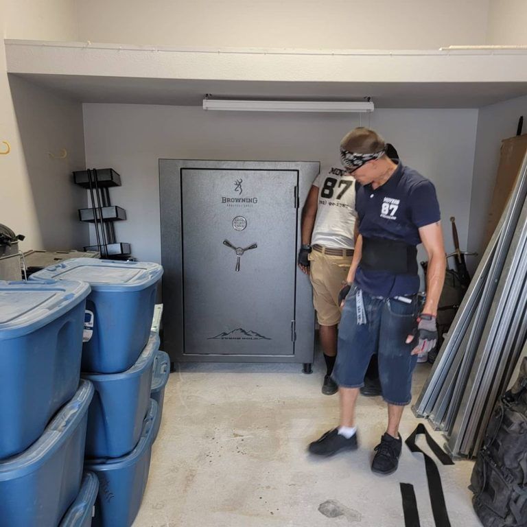 Two people moving a large, gray safe in a room with blue storage bins. One wears goggles and a back brace.