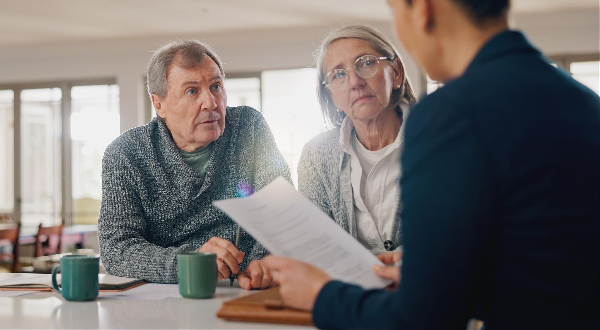 Senior couple reviews paperwork with a professional. Serious expressions. Indoor setting with sunlight.