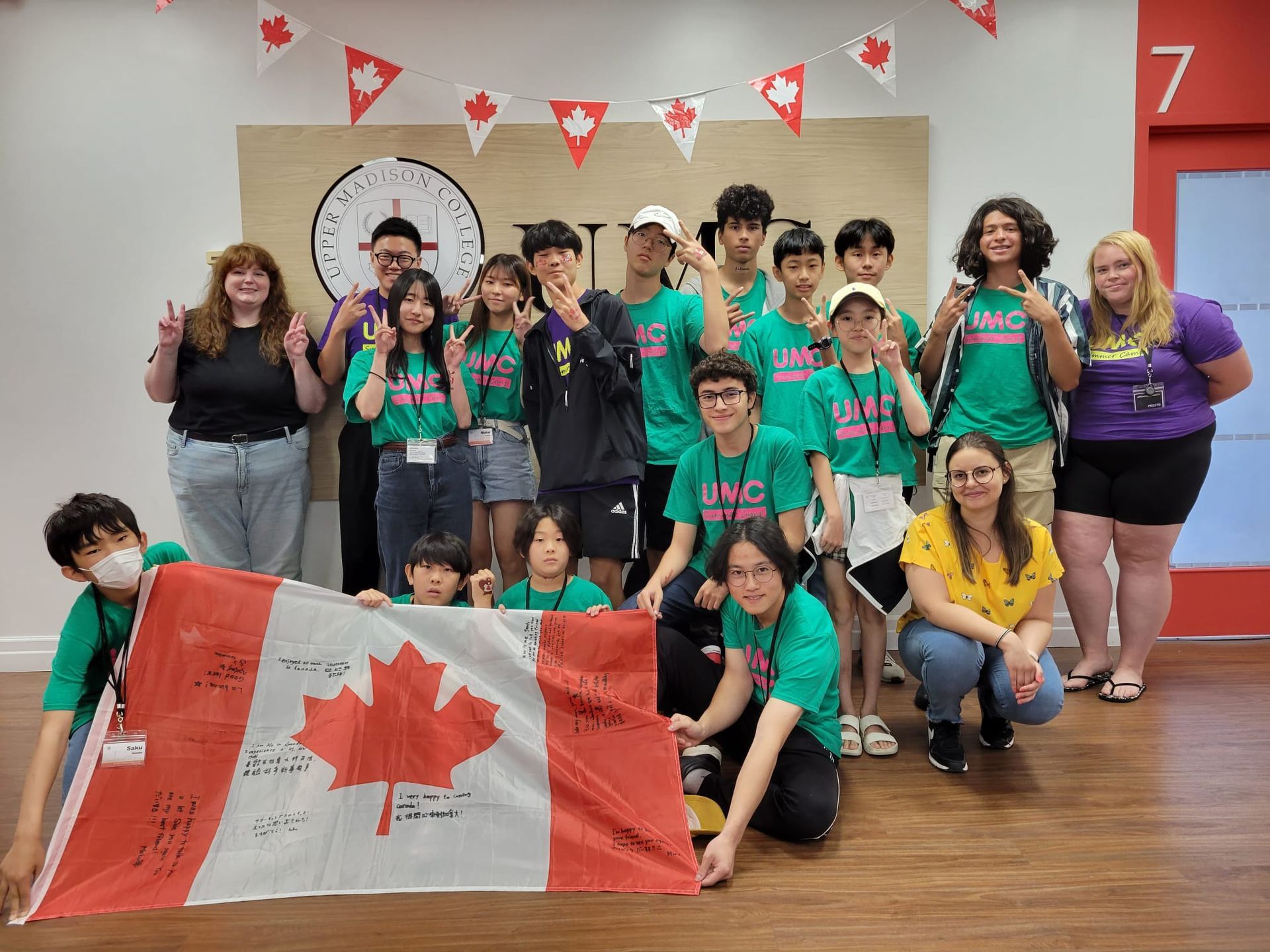 Group of people hold a Canadian flag, celebrating, wearing green shirts.  Red and white decor in room.