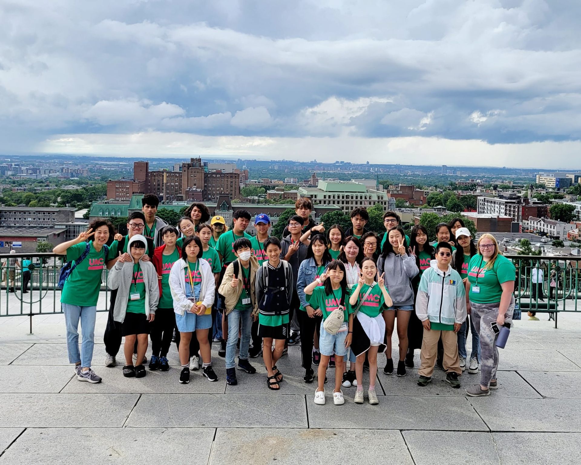 Group of people wearing green shirts posing on a city viewpoint. Overcast sky with buildings in background.