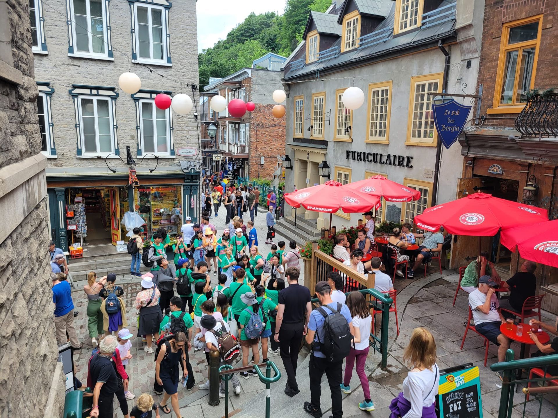Cobblestone street scene, Quebec City. People walk past shops and cafes. Group in green shirts, red umbrellas, hanging lanterns.