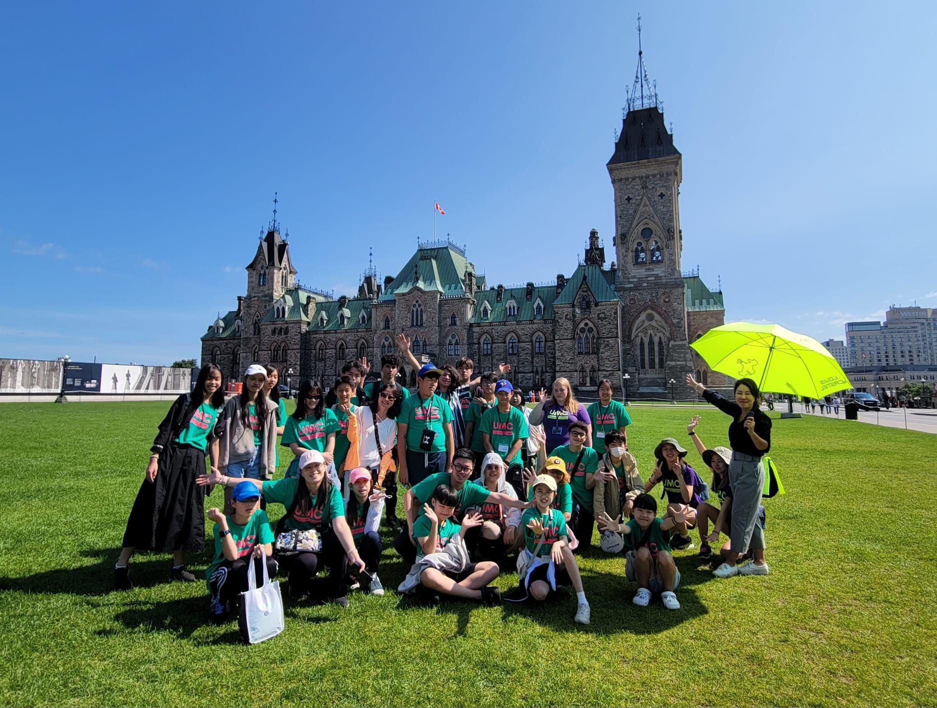 Group of people in green shirts pose on a lawn in front of the Parliament Buildings under a blue sky.