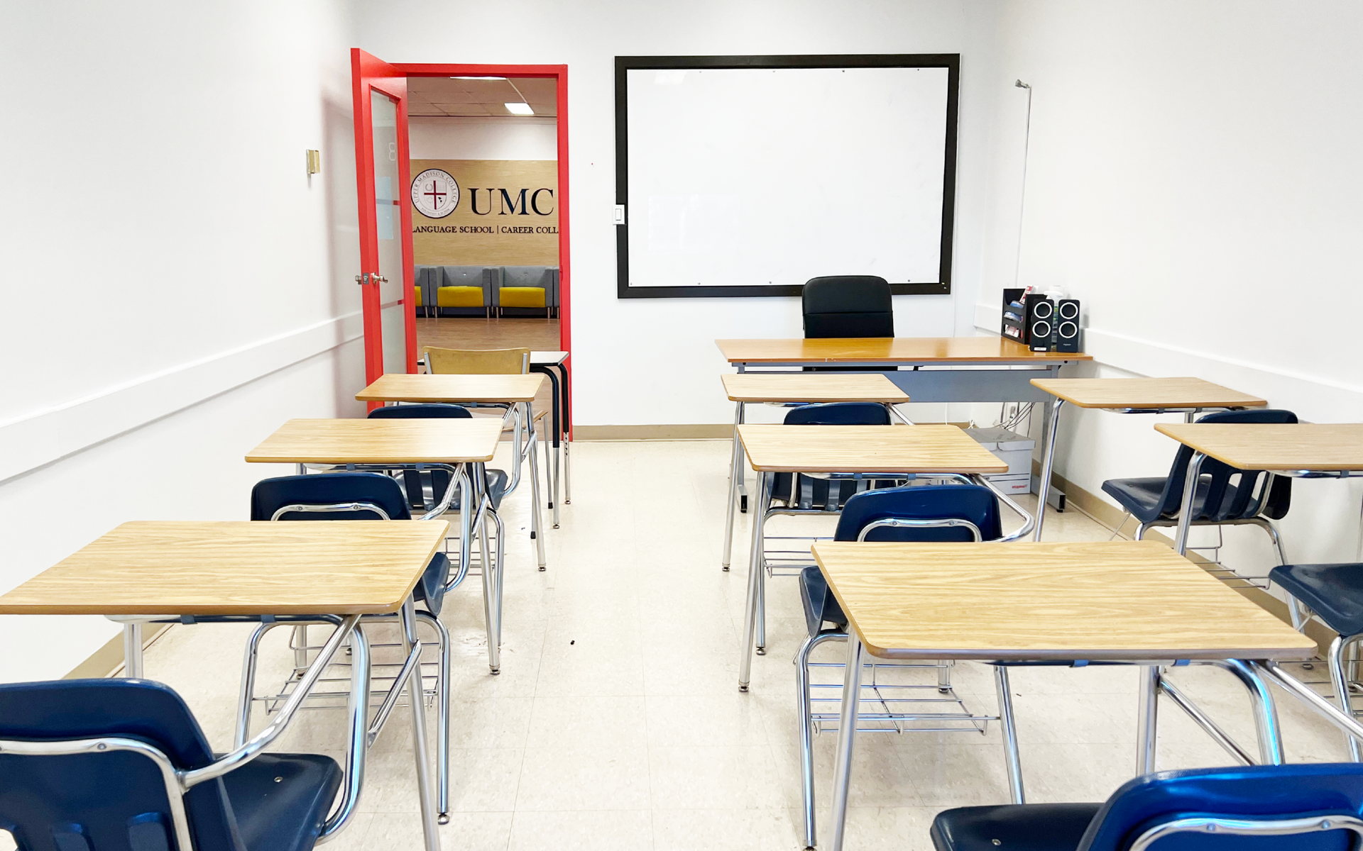 Empty classroom with desks, chairs, and projector screen. Red doorway leads to hallway.