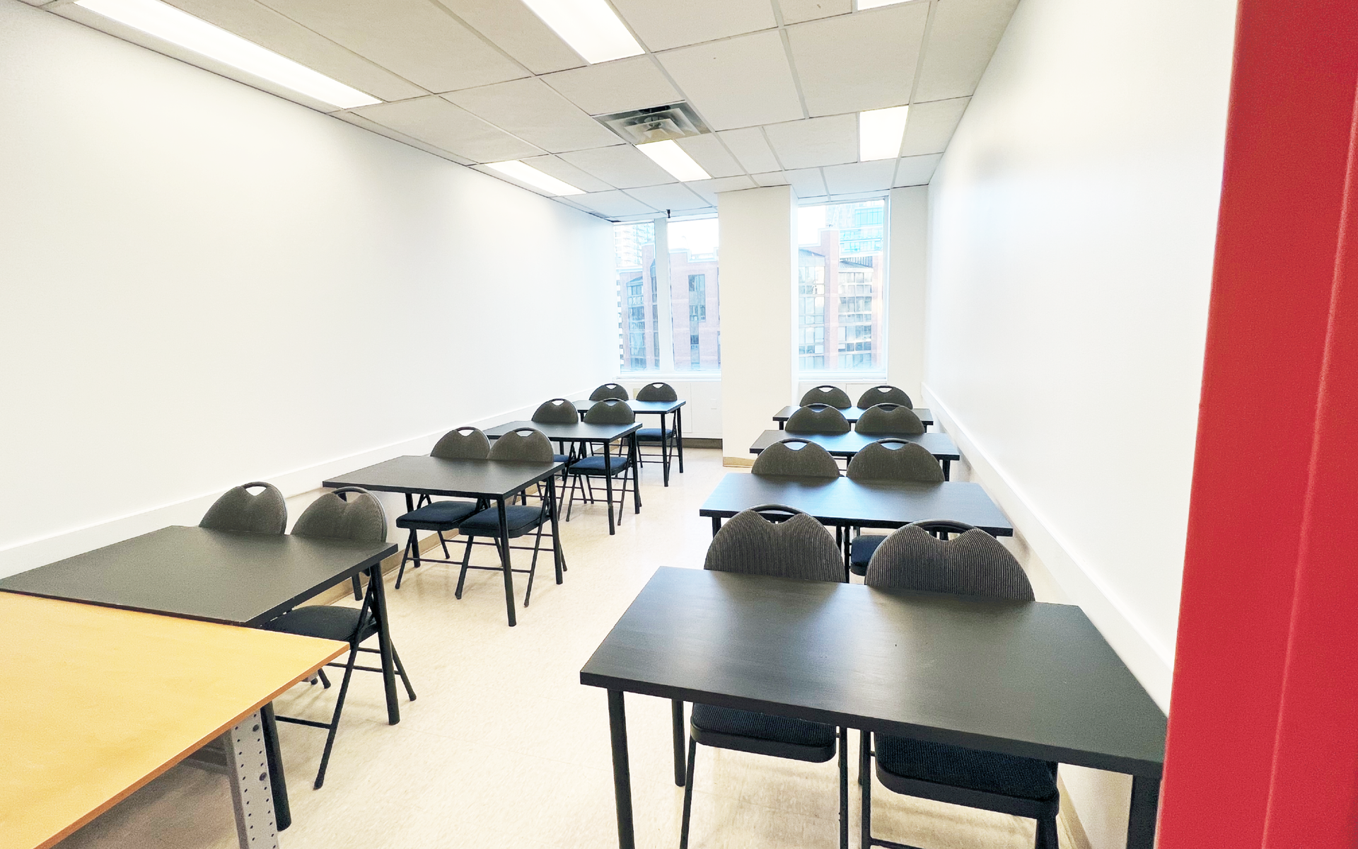 Empty classroom with tables and chairs, white walls, and bright overhead lights.