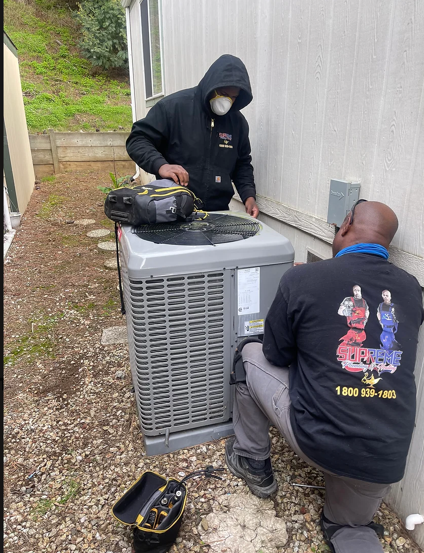 Two men are working on an air conditioner outside of a building.