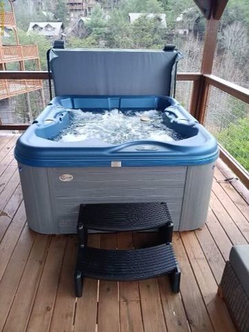 Hot tub on wooden deck, cover open. Steps in front. Scenic forest view in background.