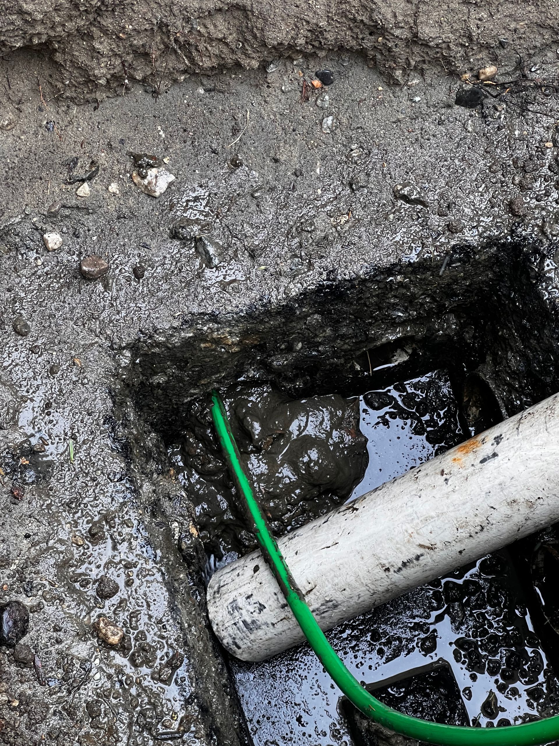 A green wire is connected to a white pipe in a hole in the ground.