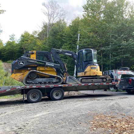 Une pelle mécanique John Deere jaune et noire chargée sur une remorque à plateau stationnée sur un terrain gravillonné près d'une forêt.