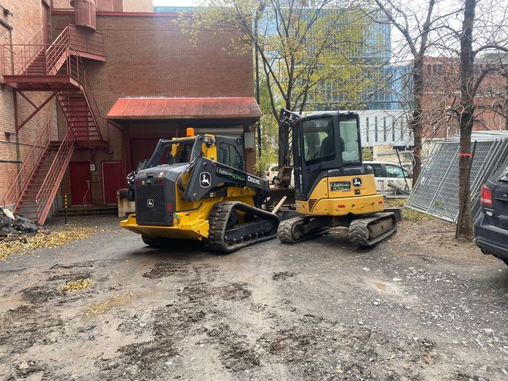 Deux engins de chantier chenillés John Deere, jaune et noir, sont stationnés sur un terrain gravillonné à côté d'un bâtiment en briques.