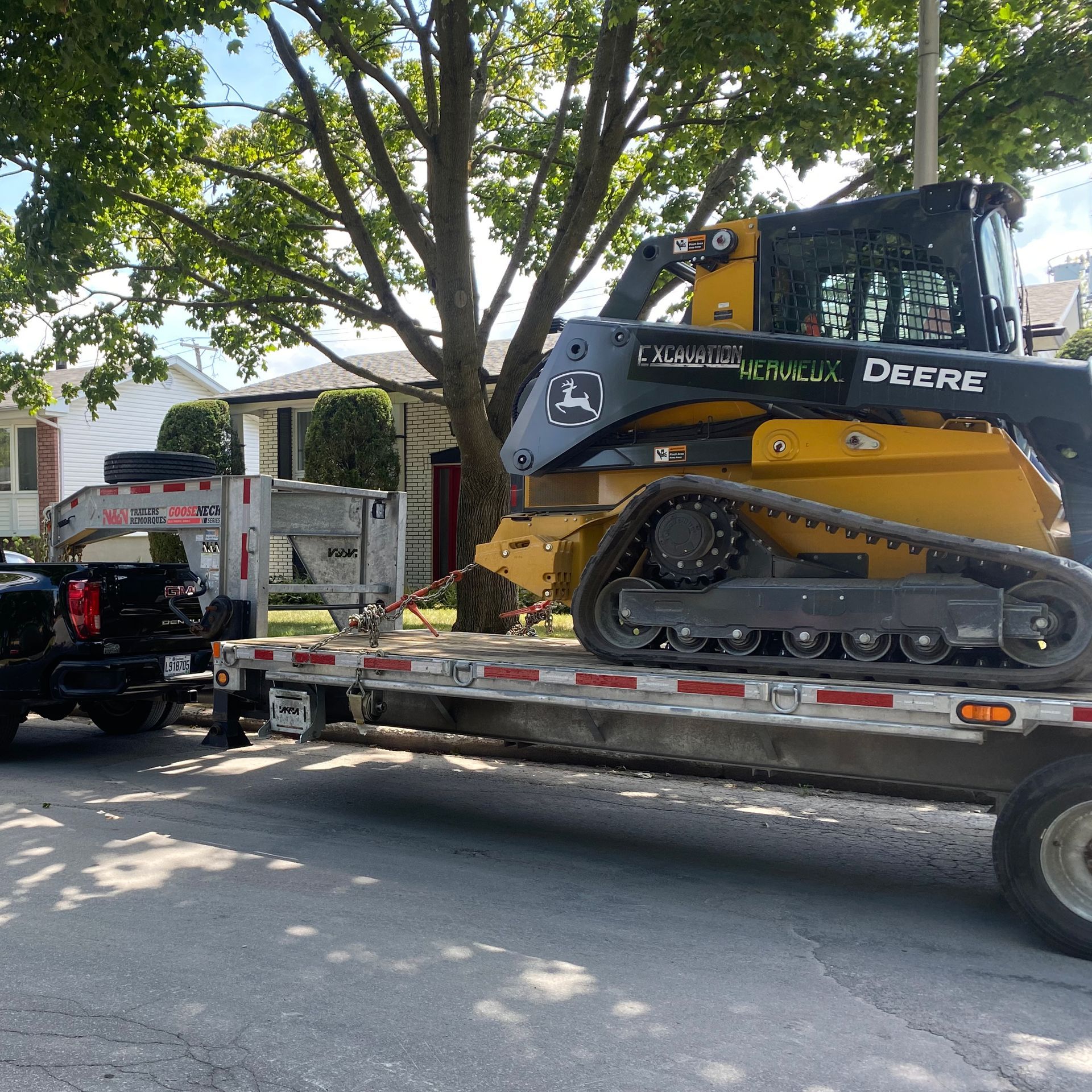 Une chargeuse compacte John Deere jaune est posée sur une remorque à plateau, tractée par une camionnette dans une rue de banlieue.