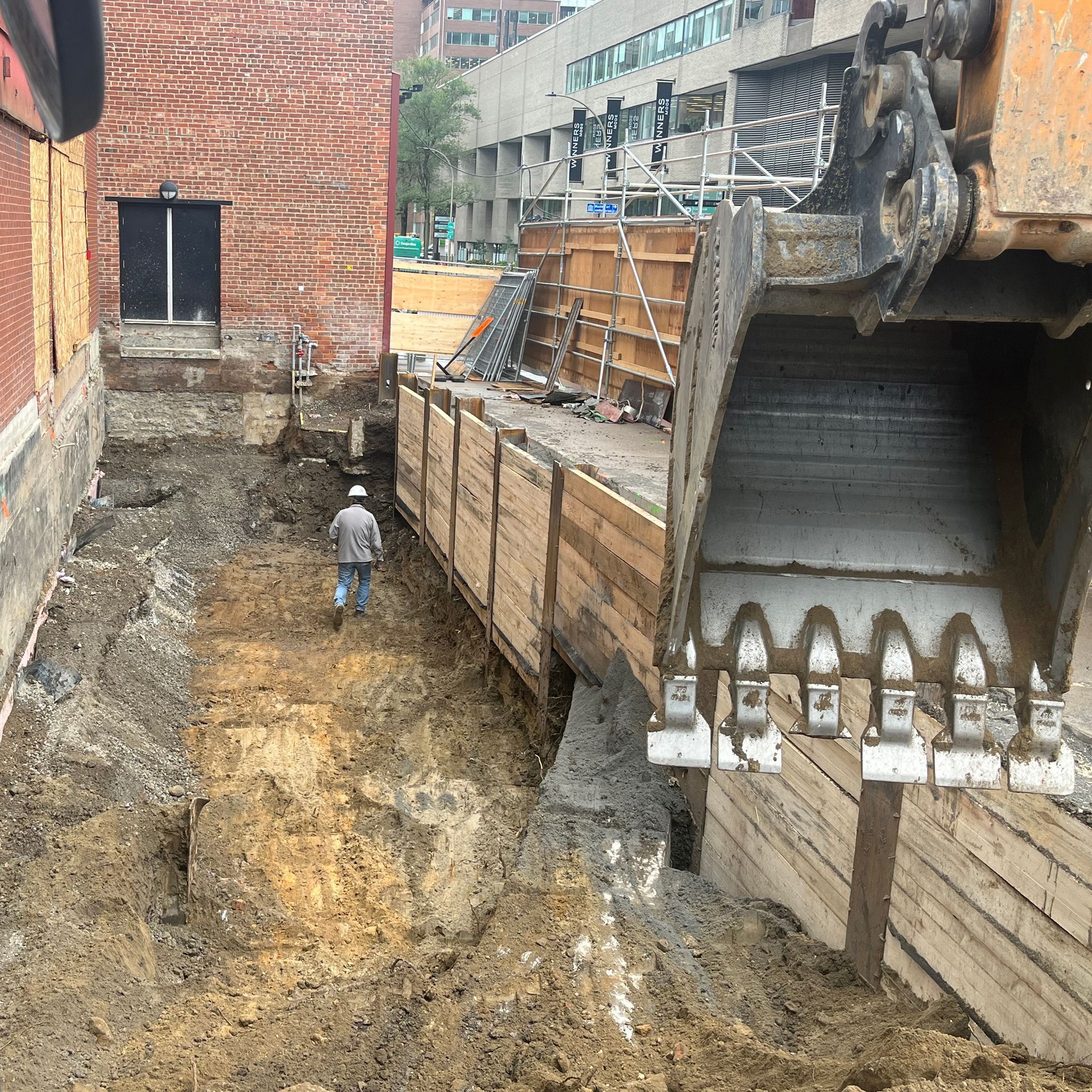 Un ouvrier du bâtiment traverse une tranchée profonde et boueuse, longeant un mur étayé, vu depuis le godet d'une excavatrice stationnée.