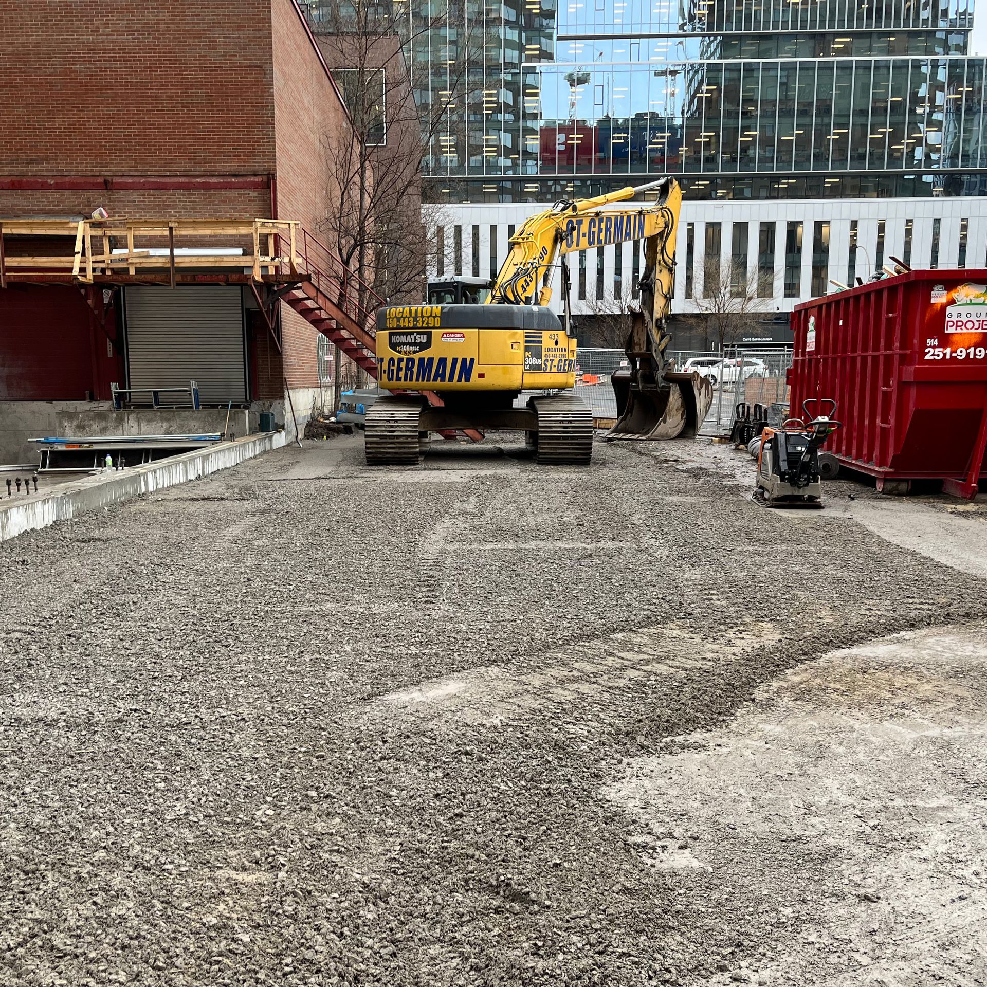 Une pelle mécanique Komatsu jaune est garée sur un chantier de gravier, à côté d'un bâtiment en briques et d'une grande benne à ordures rouge.
