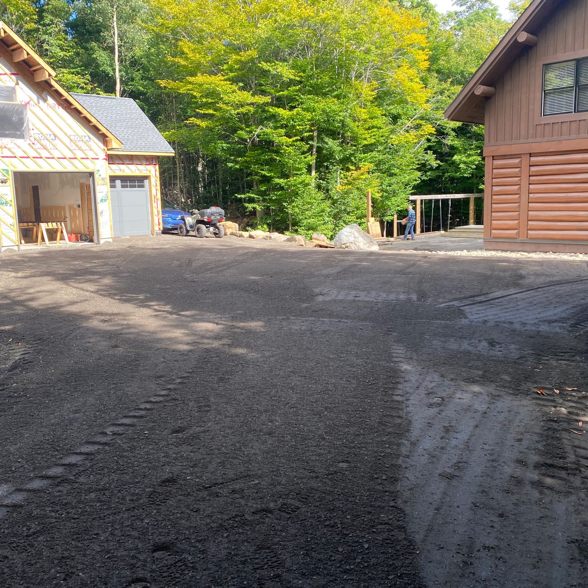 Une allée de gravier se situe entre un garage partiellement aménagé à gauche et un chalet en rondins à droite, près d'une forêt verdoyante.