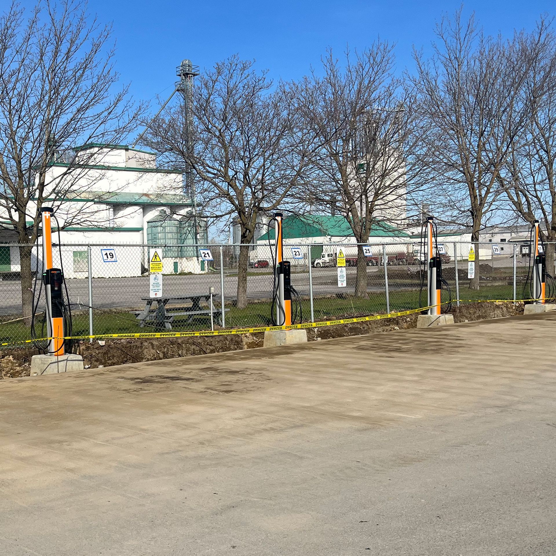 Une rangée de bornes de recharge pour véhicules électriques, orange et noires, installées sur un parking, le long d'une clôture en grillage.