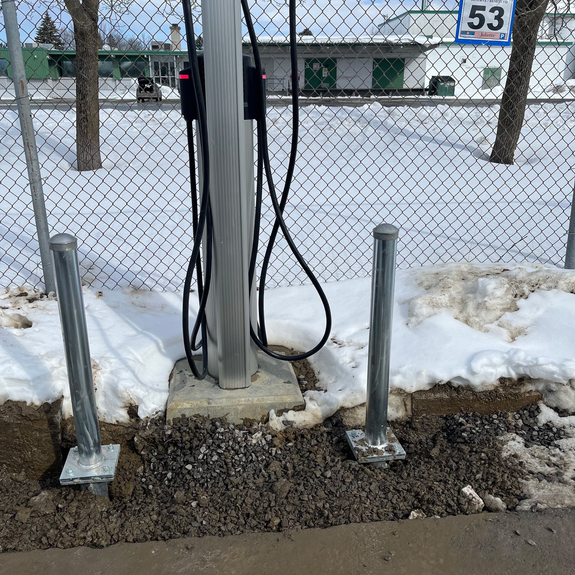 Une station de recharge pour véhicules électriques se dresse dans une zone enneigée, protégée par deux bornes métalliques devant une clôture en grillage.