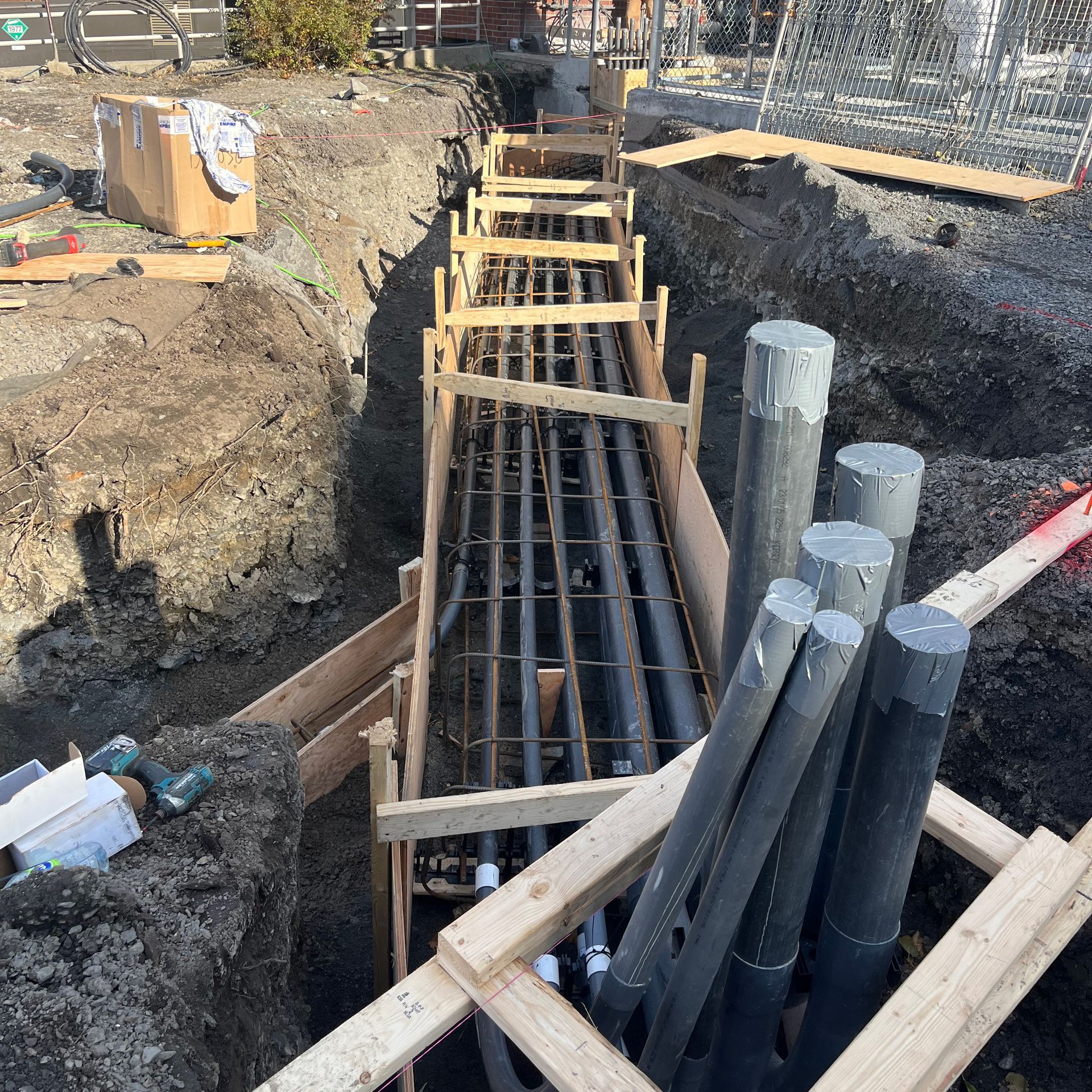 Une tranchée de construction ouverte contenant des conduits gris et une armature en acier, renforcée par un coffrage en bois.