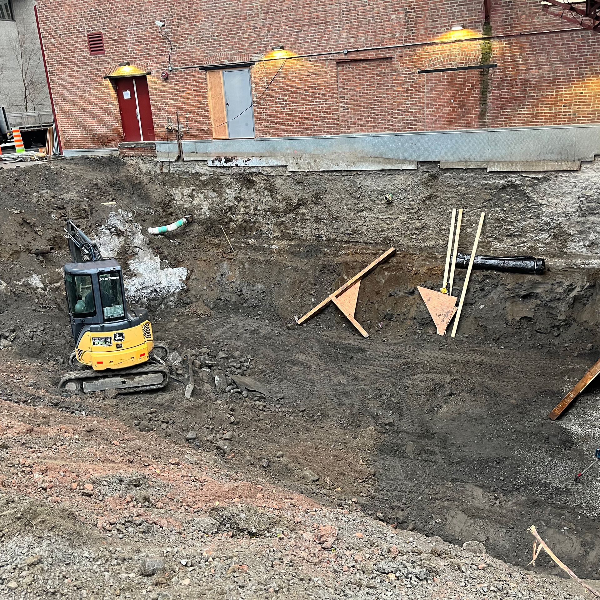 Une petite excavatrice jaune se trouve dans une fosse de terrassement près d'un bâtiment.