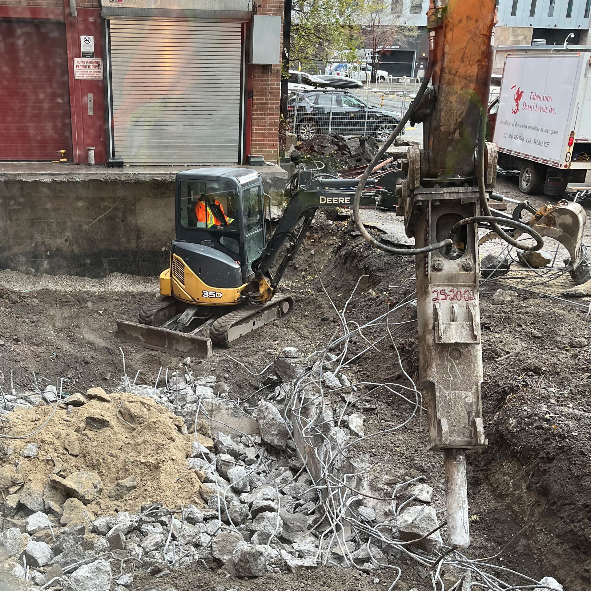 Une mini-pelle jaune et noire travaille sur un chantier de construction, avec un gros marteau hydraulique au premier plan.