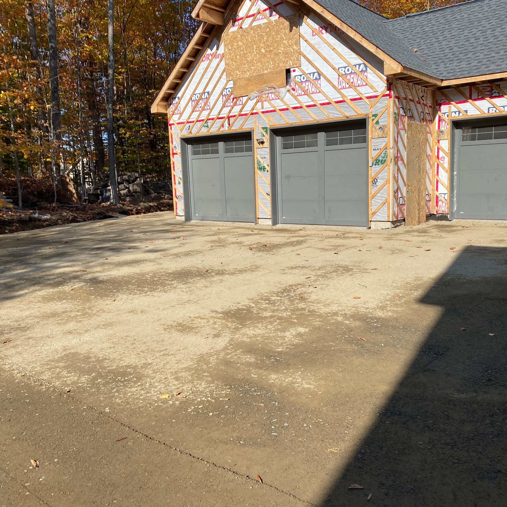 Une maison neuve en construction, avec trois portes de garage grises et une allée de gravier, dans un cadre boisé.