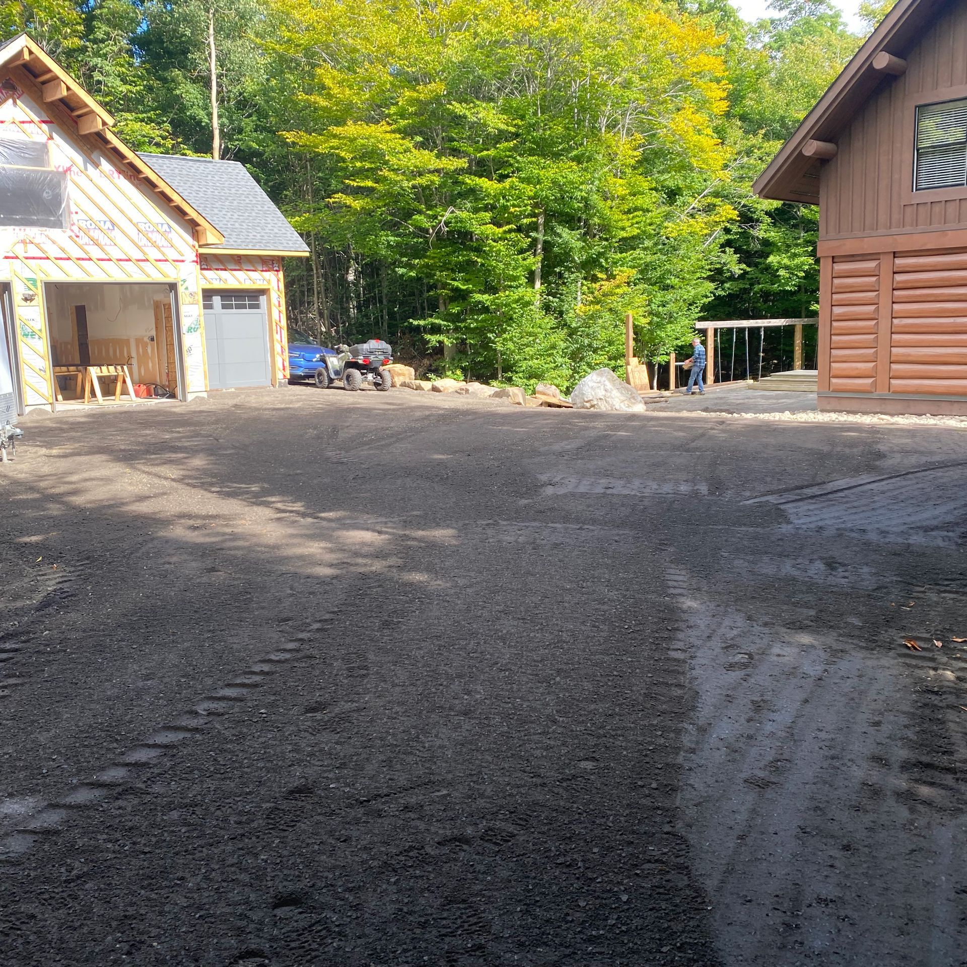 Une large allée de gravier mène à un garage moderne en construction, jouxtant un bâtiment rustique en rondins près d'un bois dense et verdoyant.