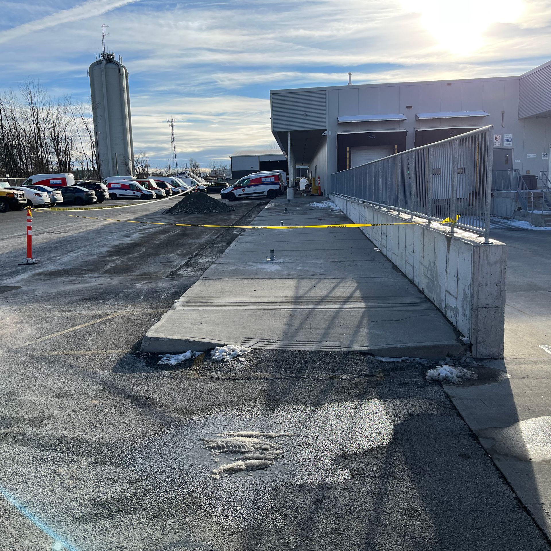 Une aire de chargement pavée à l'extérieur d'un entrepôt, bloquée par du ruban de signalisation jaune, avec des voitures garées en arrière-plan.