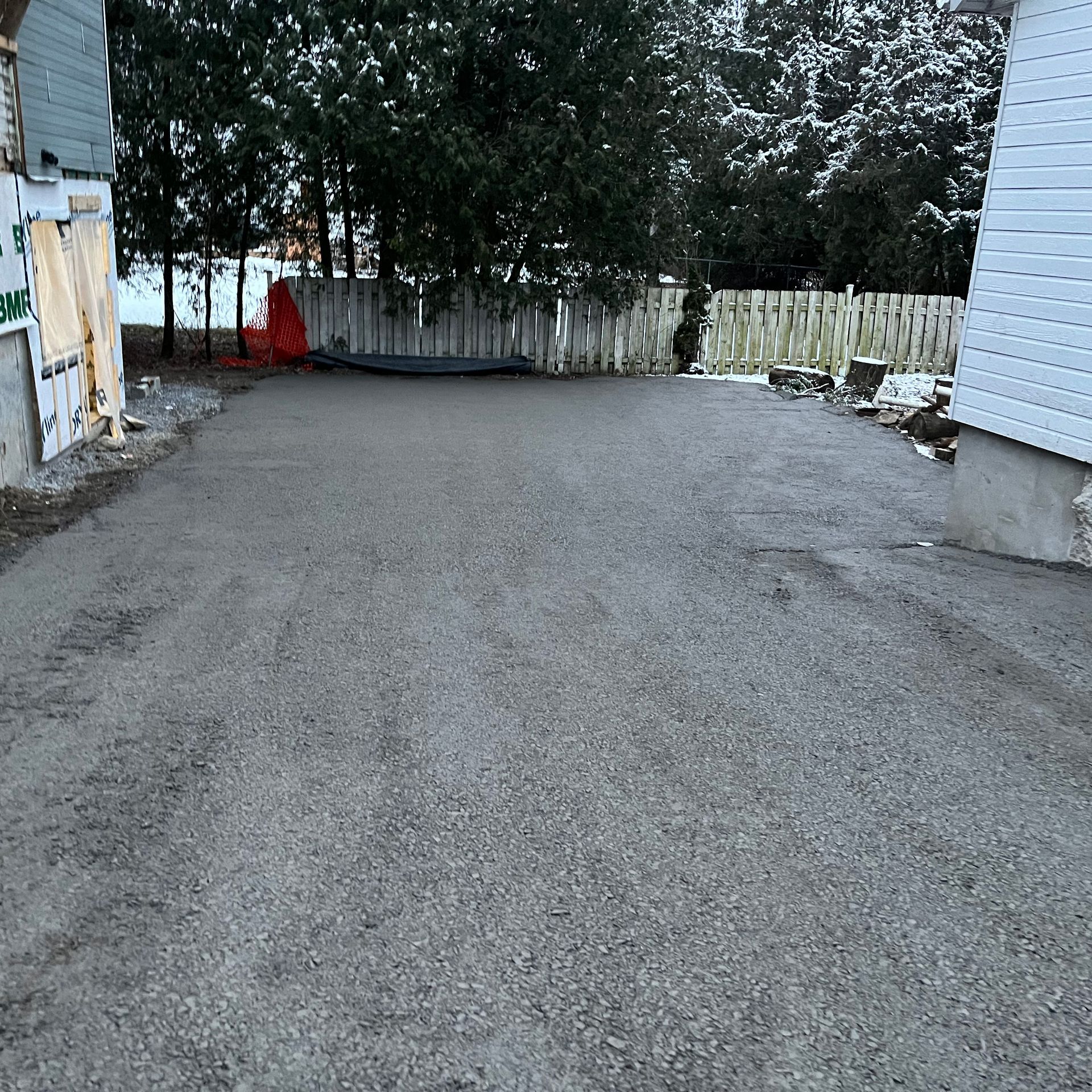 Une allée de gravier se situe entre deux murs blancs, menant vers une clôture en bois et des arbres par une journée d'hiver.