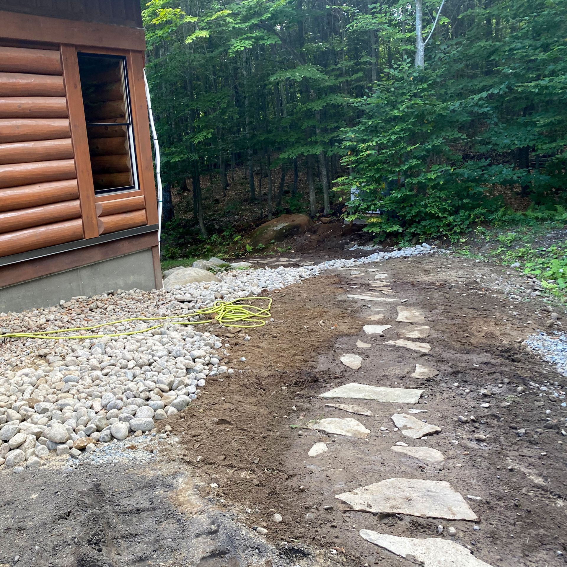 Un chemin de pierre mène à l'extérieur d'une cabane en rondins, bordée de pierres claires et d'un arrière-plan boisé.