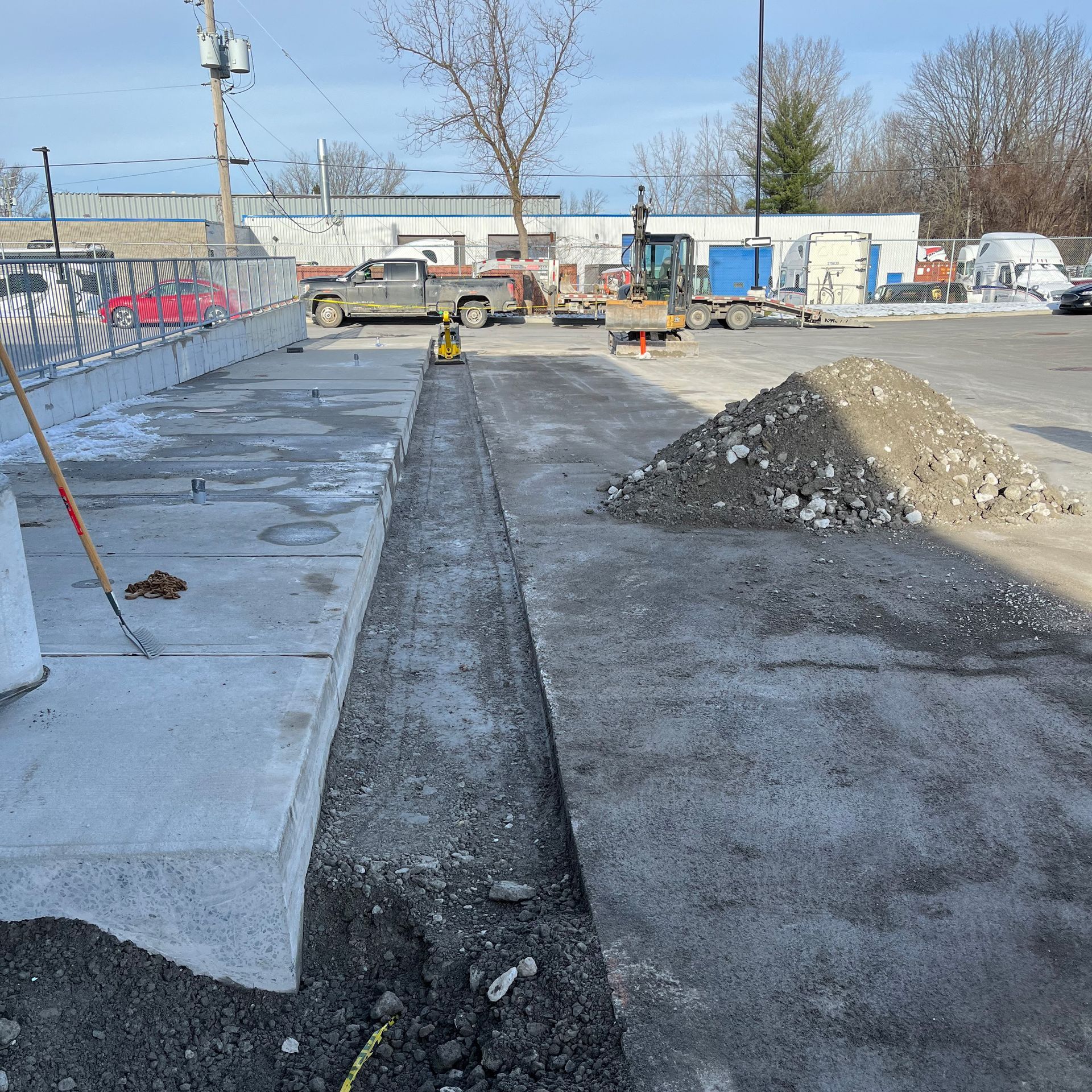 Chantier montrant une longue tranchée creusée entre un trottoir en béton et une zone de gravier non pavée.