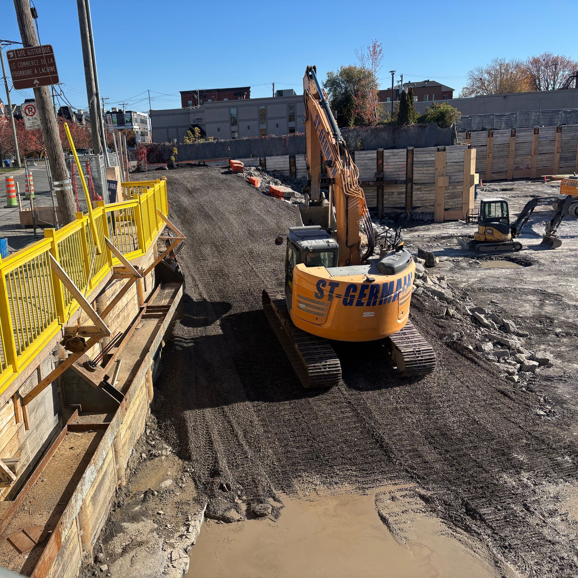 Une pelleteuse jaune sur un chantier de construction avec des murs de soutènement en bois, une clôture de sécurité jaune et une fosse boueuse.