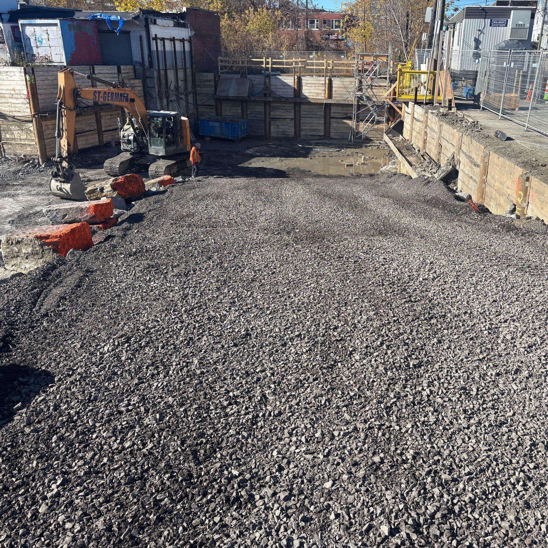 Une excavatrice est stationnée sur un chantier de construction rempli de gravier meuble, entouré de murs de soutènement en bois et d'équipements.