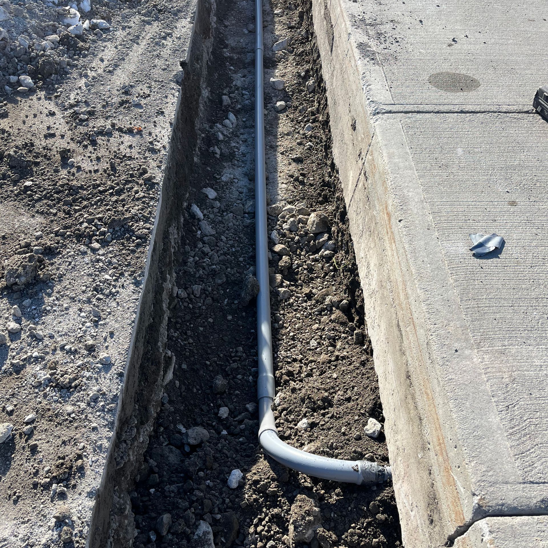 Un tuyau de conduit en PVC gris repose dans une étroite tranchée de terre, le long d'un trottoir ou d'une bordure en béton.