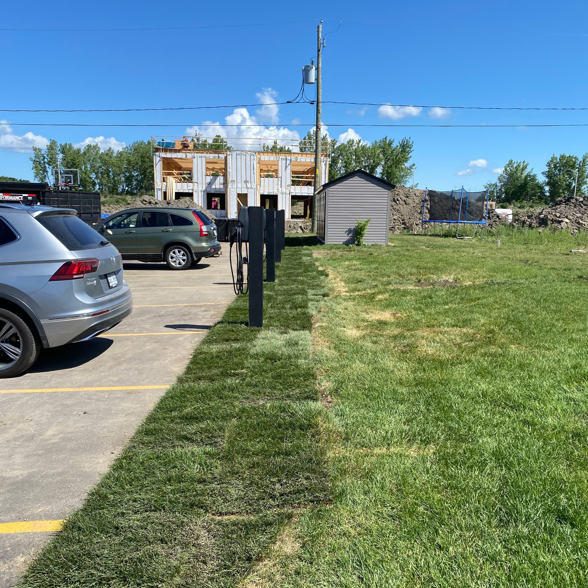 Une scène extérieure ensoleillée montre un parking avec deux voitures à côté d'une rangée de gazon nouvellement installée bordant un champ herbeux.