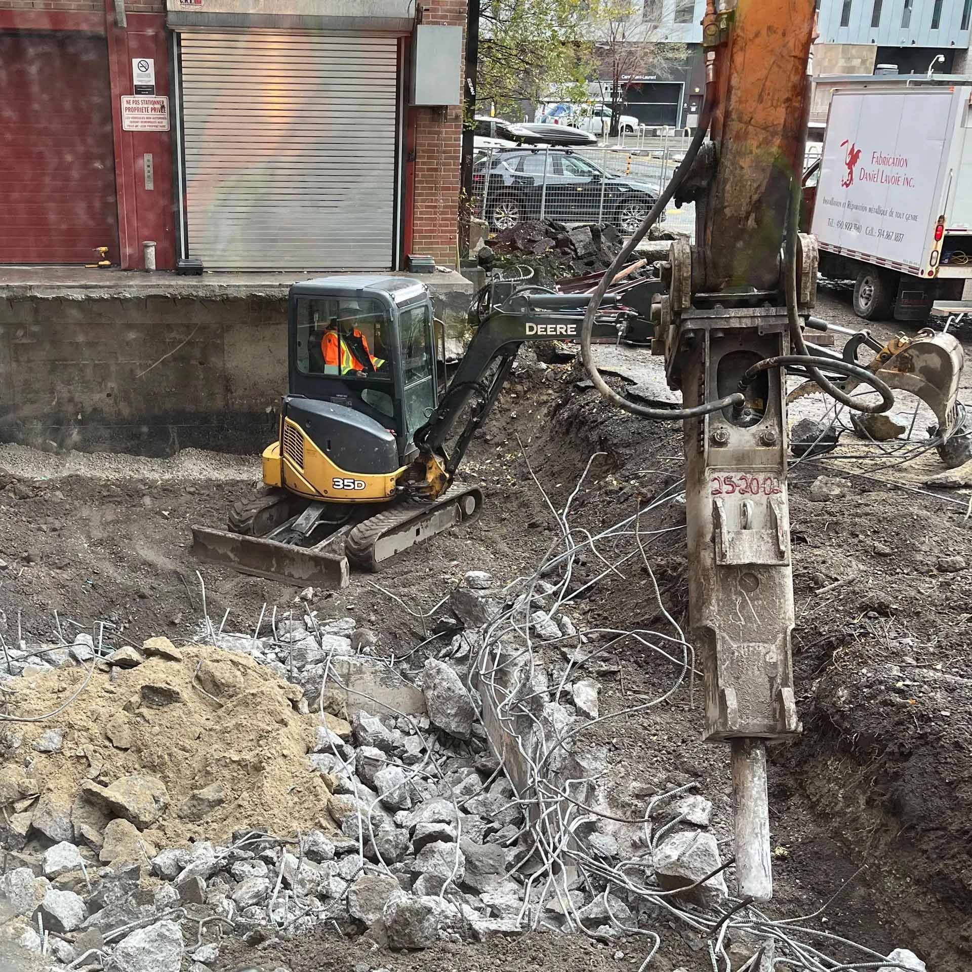 Une petite excavatrice et un marteau hydraulique, fixés à une machine plus imposante, travaillent à la démolition du béton sur un chantier de construction.