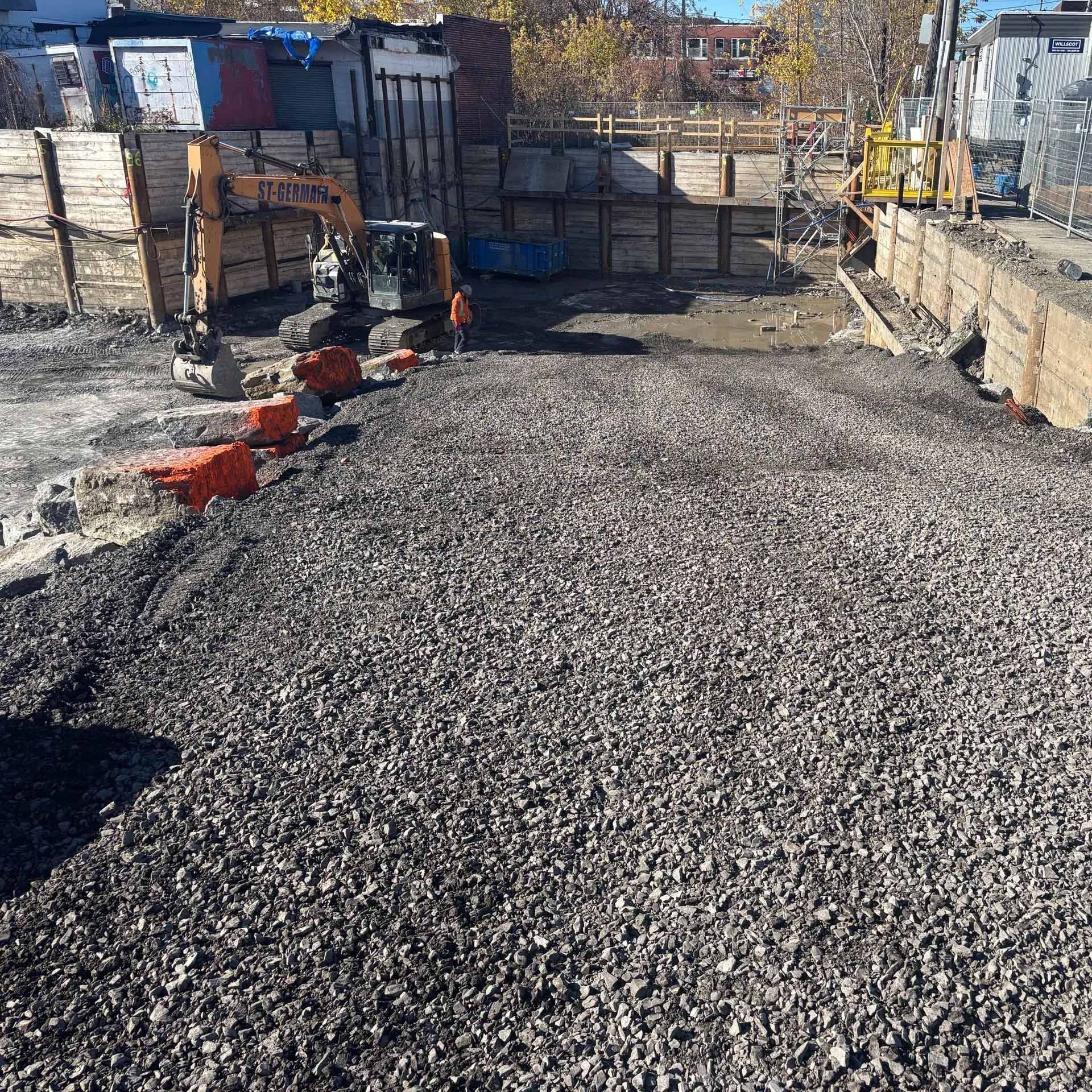 Une pelleteuse orange est posée sur un chantier en pente recouvert de gravier, avec des murs de soutènement en bois en arrière-plan.