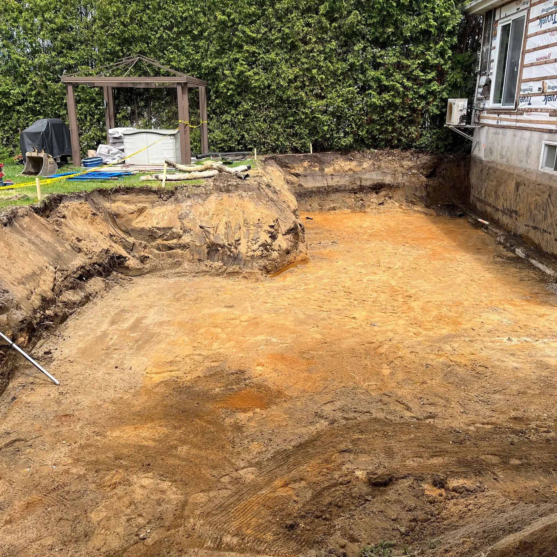 Un jardin en terre battue jouxte une maison aux fondations apparentes, avec une treille en bois en arrière-plan.