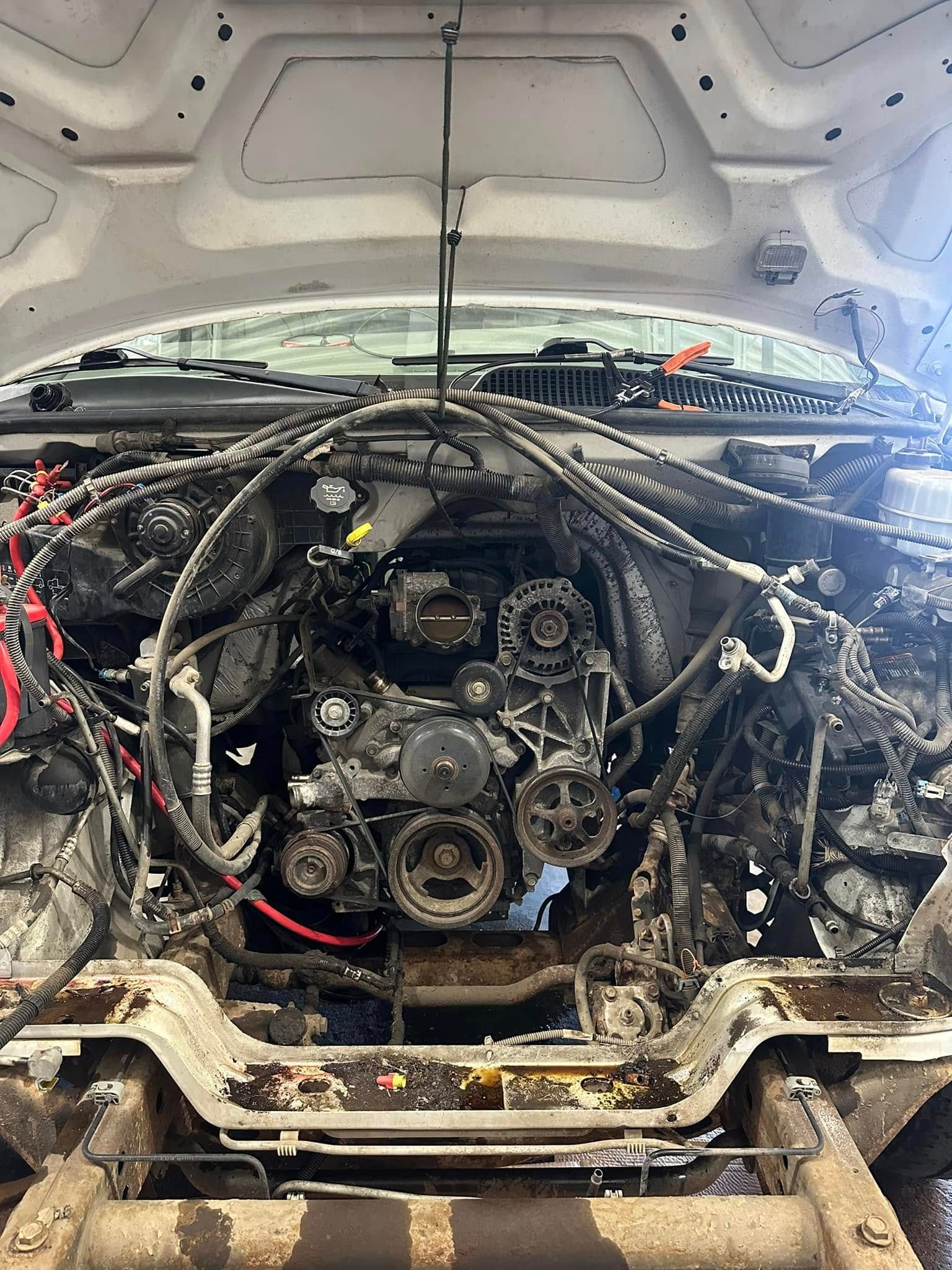 Car engine bay with exposed hoses, pulleys, and wiring under an open hood, showing heavy grime and damage | MRM Auto LLC