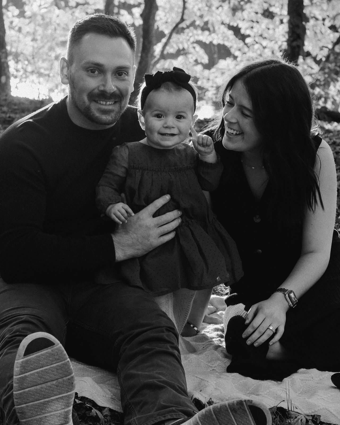 Smiling family of three sitting outdoors, holding a baby in front of a flowering tree | MRM Auto LLC