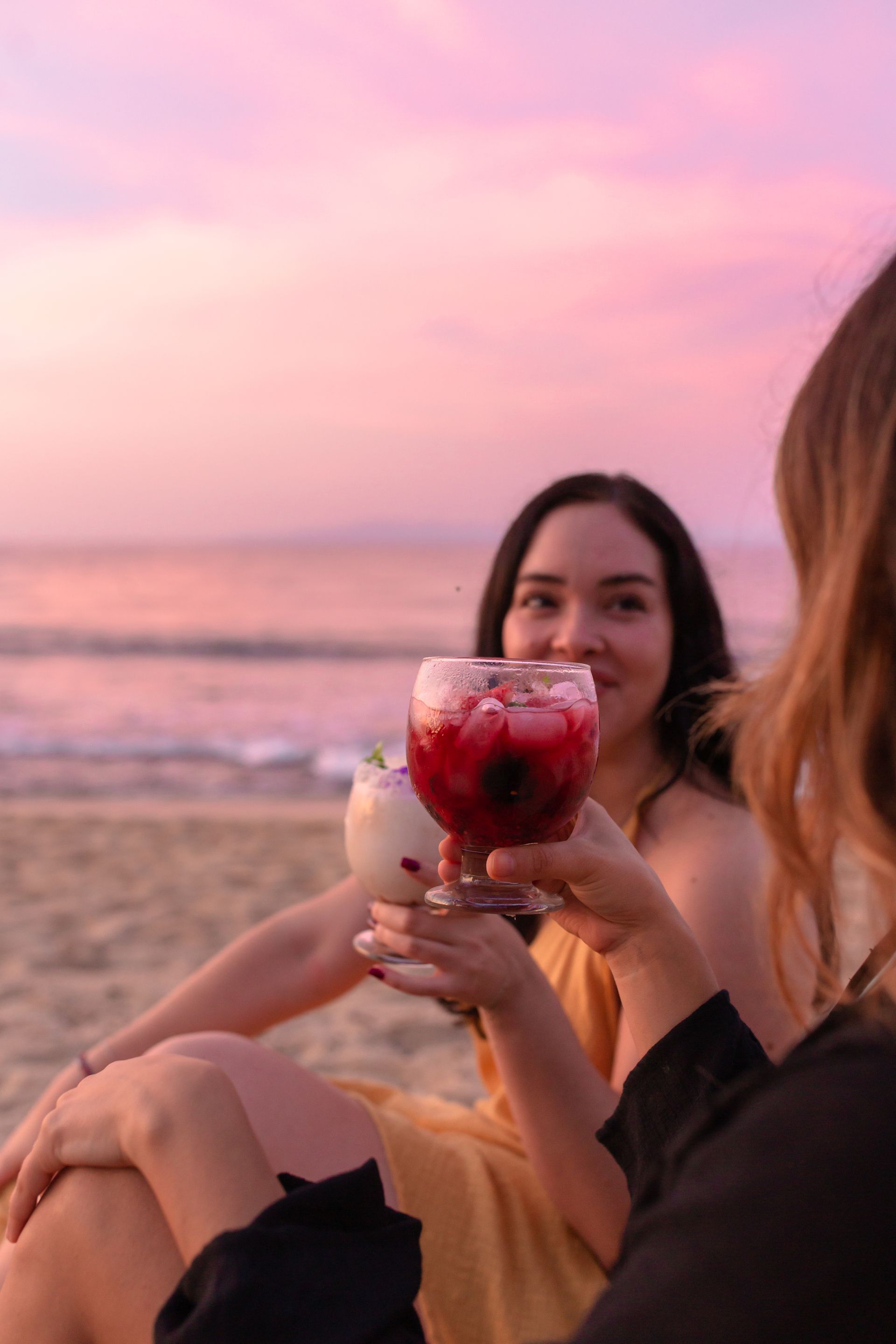 Dos mujeres están sentadas en la playa sosteniendo bebidas en sus manos.