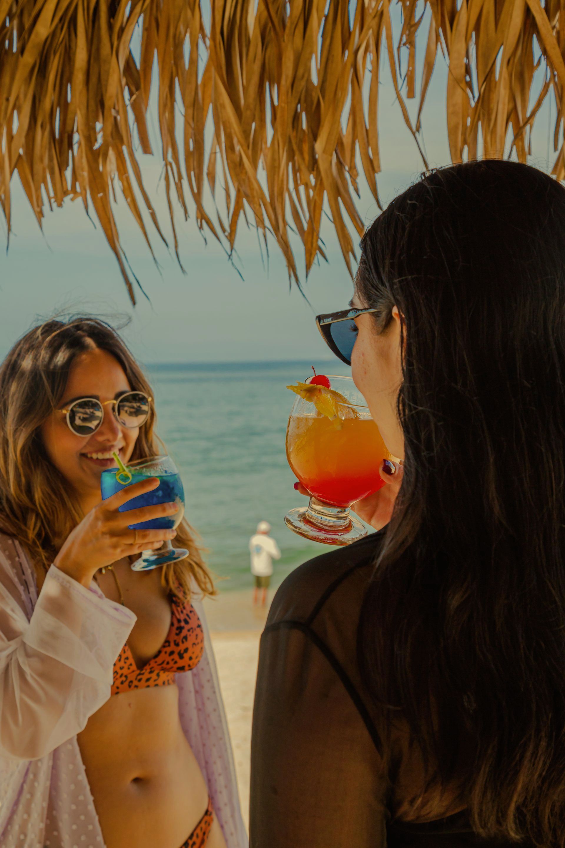 Dos mujeres están bebiendo cócteles en la playa bajo un techo de paja.