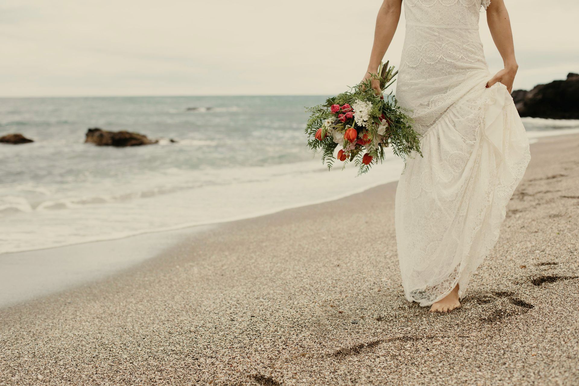 Una mujer con un vestido de novia sostiene un ramo de flores en la playa.