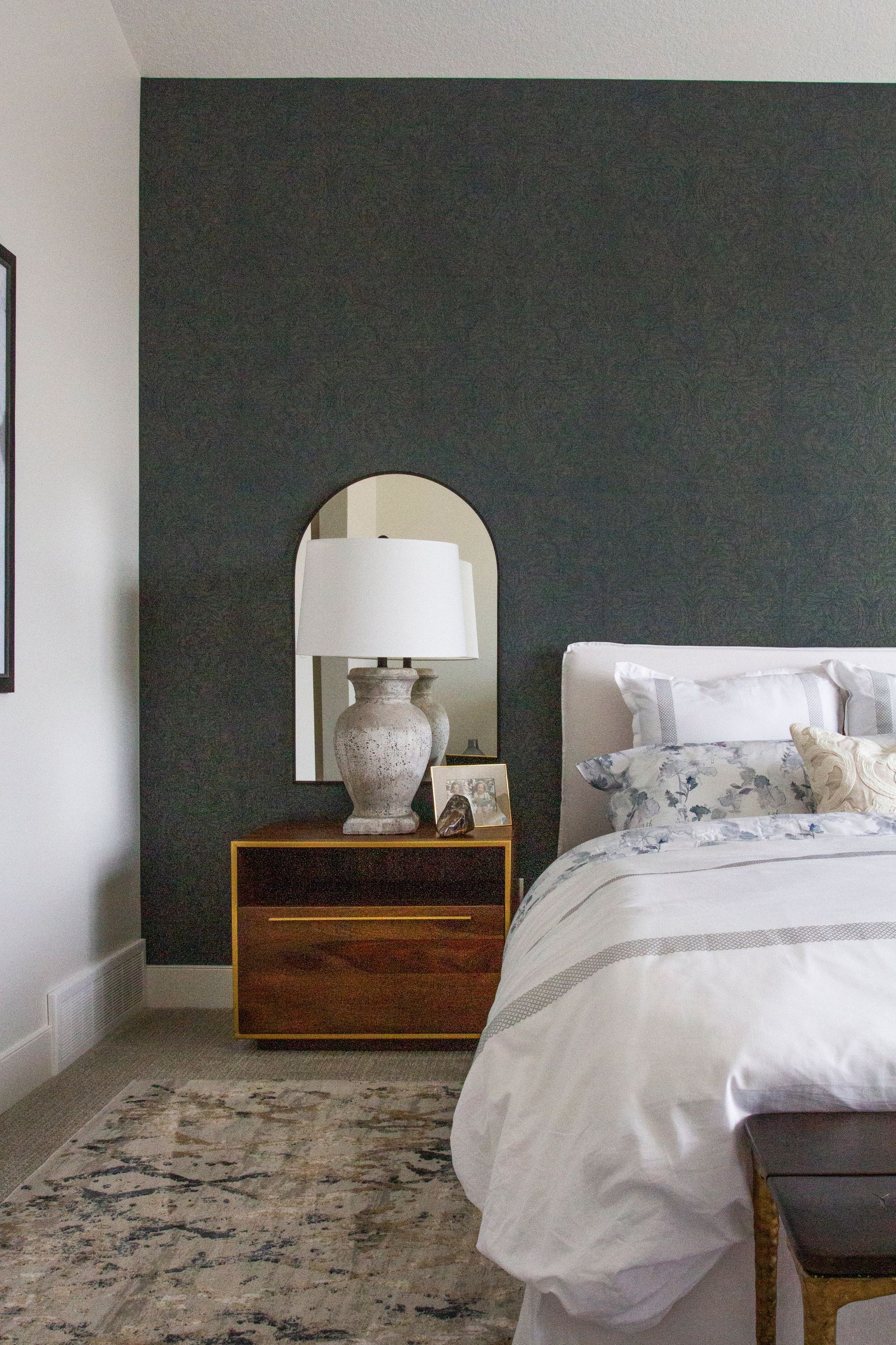 A bedroom with a bed , nightstand , lamp and mirror.