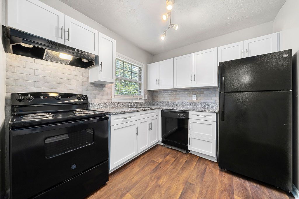Bright white kitchen with black appliances, wood floors, and a window over the sink